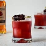pink drink with three cherries on top and bottle of bourbon on the left side