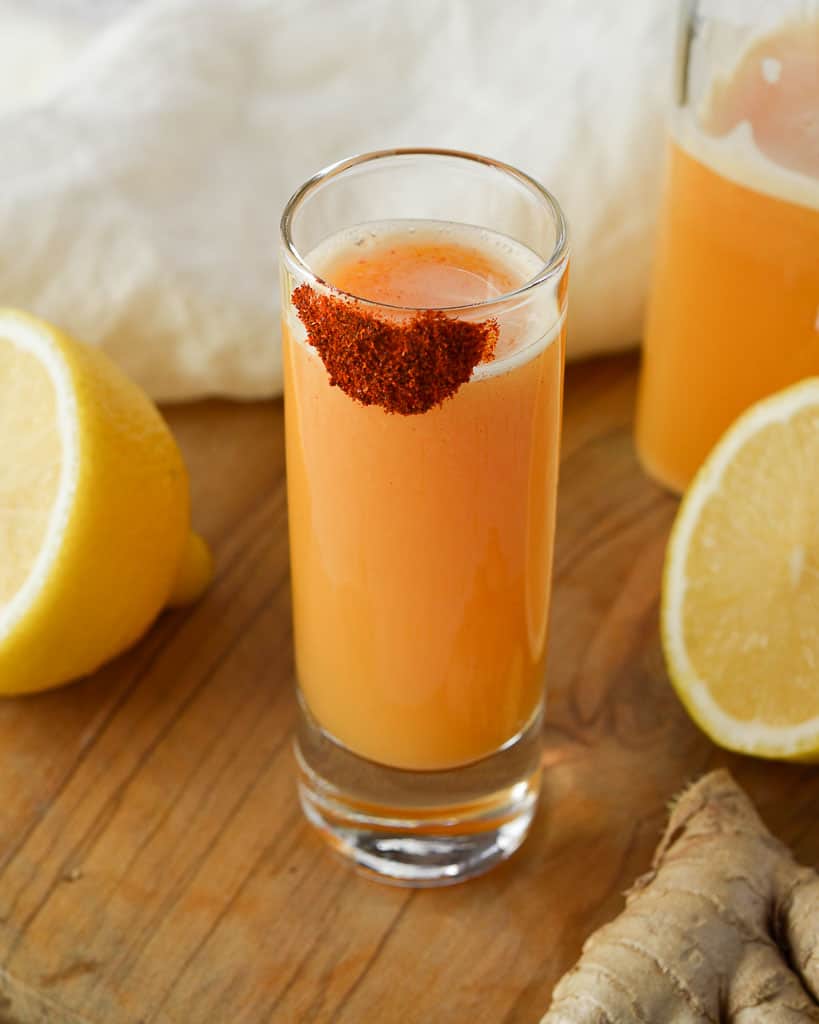 a lemon ginger cayenne shot with cayene powder on the rim and lemons in the background