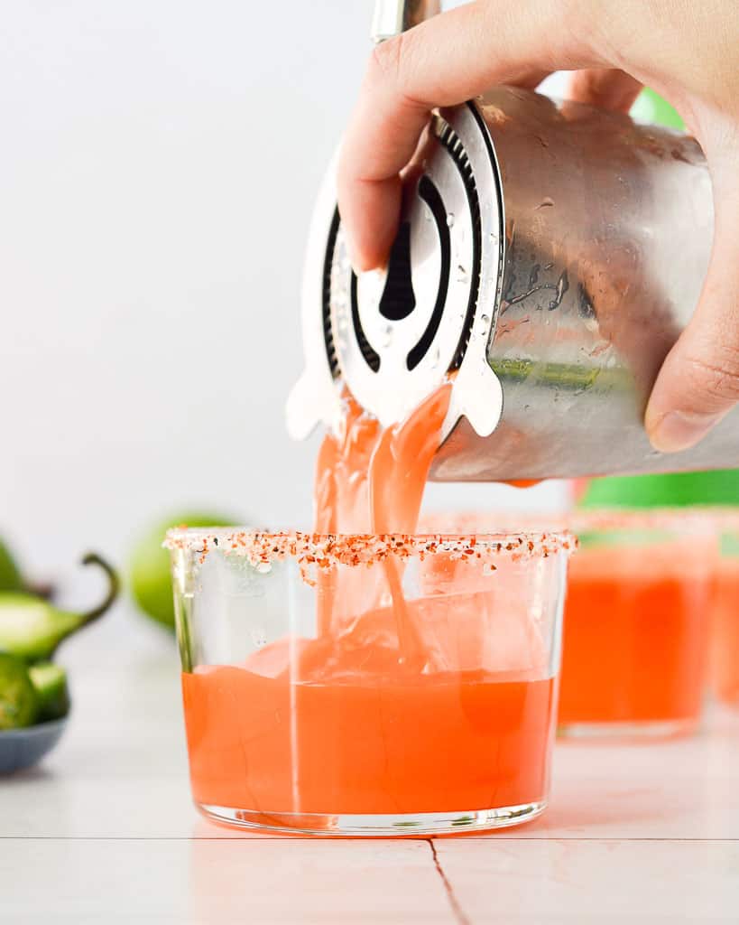pouring a margarita carrot juice cocktail from a cocktail shaker into a short glass.