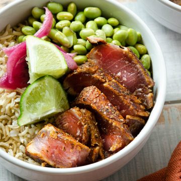 a white bowl with slices of seared ahi tuna, limes, pickled red onions and edamame over brown rice