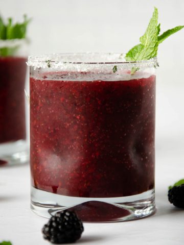 small glass filled with a frozen blackberry margarita with a mint leaf on top. frozen blackberry glass in the back left