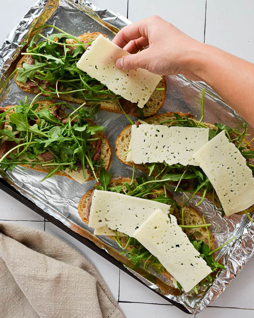 a hand adding a slice of cheese on top of an arugula sandwich