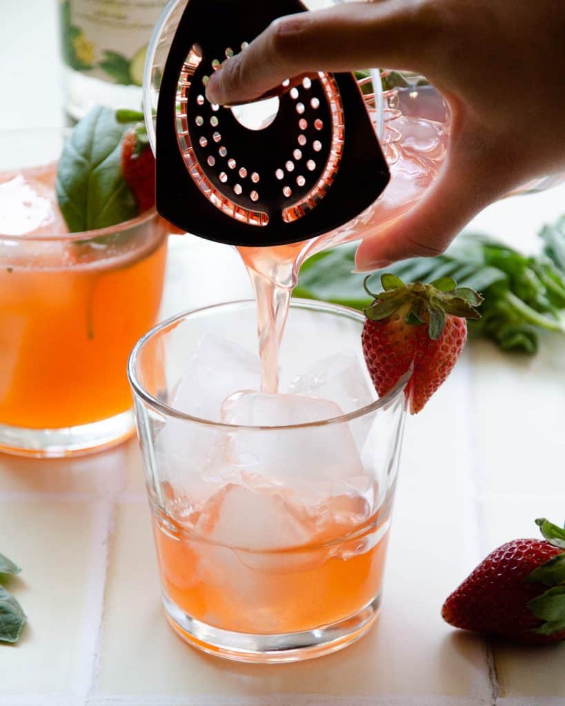 straining the cocktail into a short glass filed with ice with a strawberry garnish
