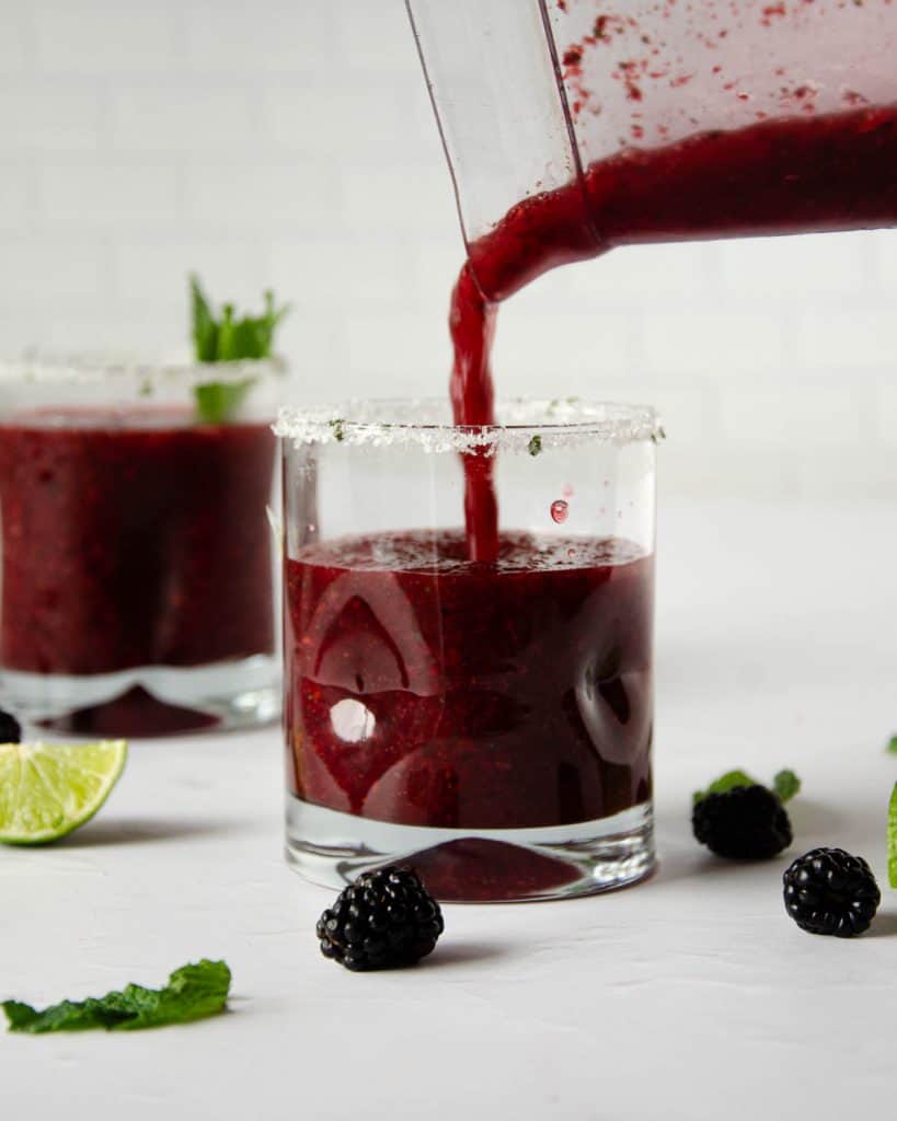 pouring frozen blackberry margarita into a half filled glass with a full glass behind to the left.