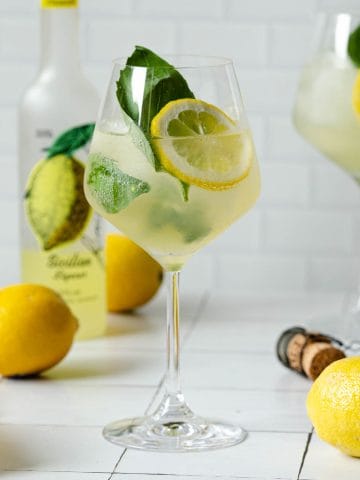 straight on photo of a limoncello spritz with a bottle of limoncello in the back left and lemons