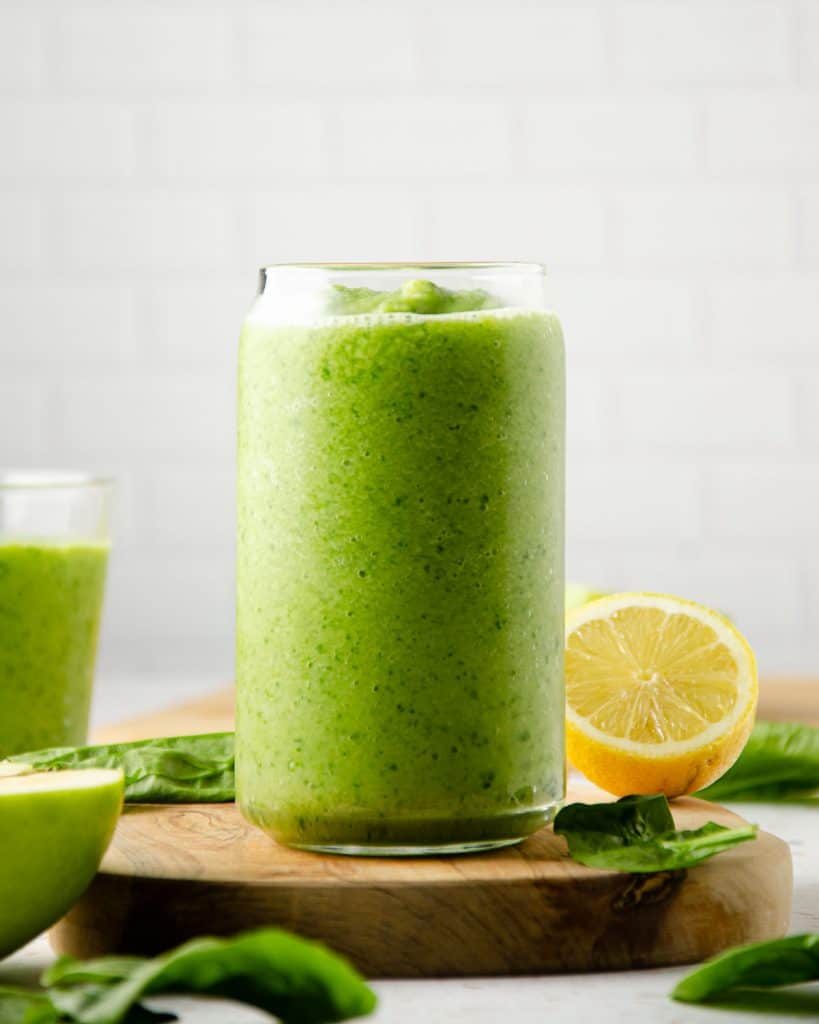 a tall green smoothie on a wooden board with lemons, green apples and spinach around