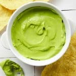 a bowl of whipped avocado surrounded by chips and a spoon of green creamy sauce