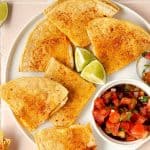square image of overhead of complete quesadilla on a white plate with a bowl of pico de gallo and limes on top. lime wedges in the corner and a spoon of pico de gallo on the side.