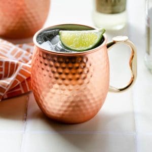 a copper mug filled with a mexican mule and garnishes with a lime wedge with another mexican mule in the background