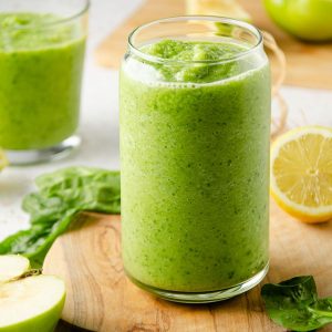 a tall green smoothie on a wooden board with lemons, green apples and spinach around