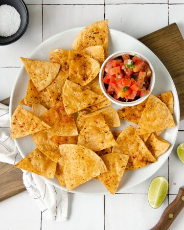 an overhead photo of a white plate topped with chili lime tortilla chips and a bowl of pico de gallo with limes scattered