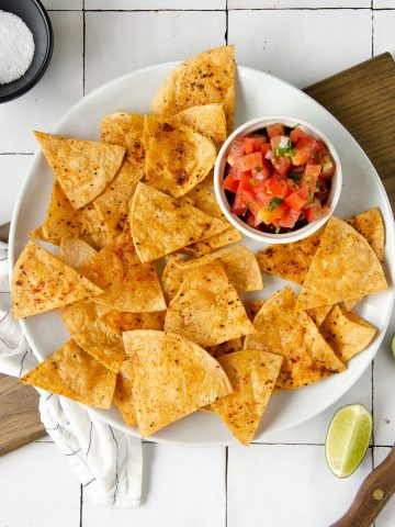 an overhead photo of a white plate topped with chili lime tortilla chips and a bowl of pico de gallo with limes scattered