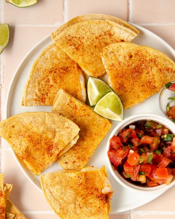 overhead of complete quesadilla on a white plate with a bowl of pico de gallo and limes on top. lime wedges in the corner and a spoon of pico de gallo on the side.