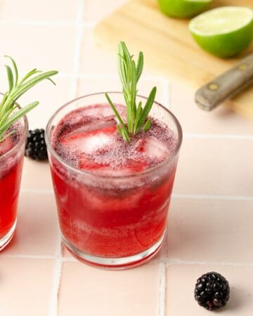 a glass of blackberry rosemary margarita garnished with a rosemary sprig and a cutting board with a fresh lime in the back right