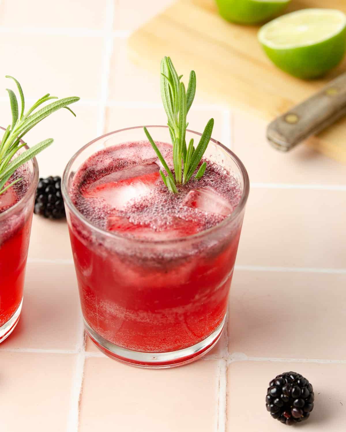 a glass of blackberry rosemary margarita garnished with a rosemary sprig and a cutting board with a fresh lime in the back right