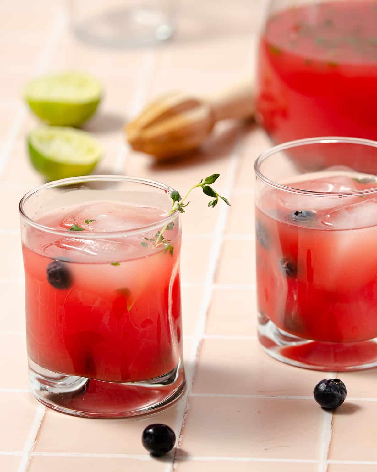 two glasses of watermelon blueberry vodka smash in front with two stray blueberries. A large pitcher to the side with two fresh squeezed limes to the back left.