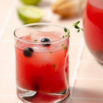 a close up of a glass of watermelon vodka punch with some blueberries and thyme decorating the glass. A large pitcher to the back right and two squeezed limes to the left