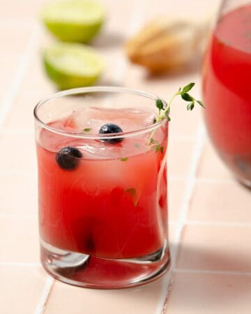 a close up of a glass of watermelon vodka punch with some blueberries and thyme decorating the glass. A large pitcher to the back right and two squeezed limes to the left