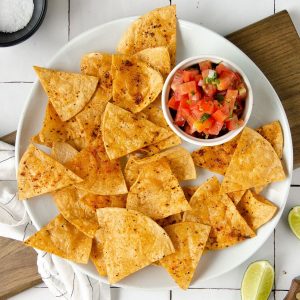square image of an overhead photo of a white plate topped with chili lime tortilla chips and a bowl of pico de gallo with limes scattered