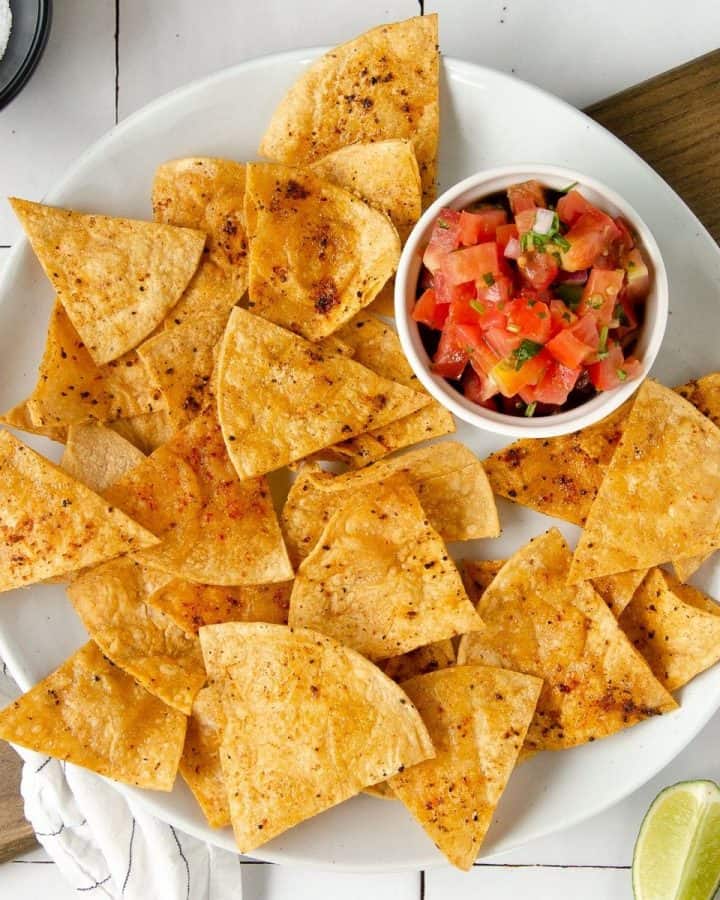 square image of an overhead photo of a white plate topped with chili lime tortilla chips and a bowl of pico de gallo with limes scattered