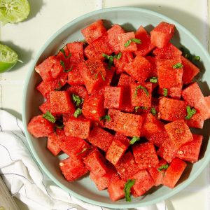 square photo of overhead view of a blue plate with watermelon tajin salad, some limes, a wooden knife and beers in the background