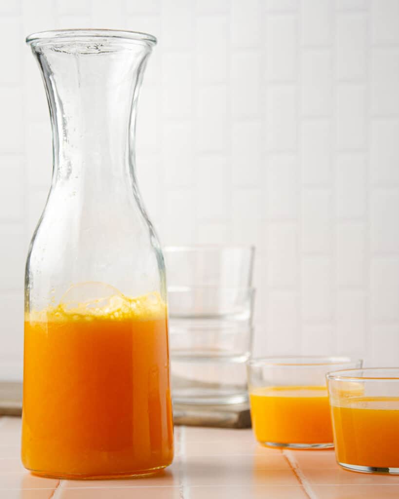 a straight on photo of a large pitcher of homemade mandarin juice with two small glasses on the right side