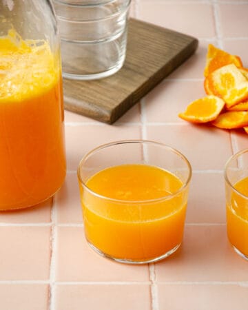 two small glasses of homemade mandarin juice with a pitcher of mandarin juice to the left side and mandarin peels in the back