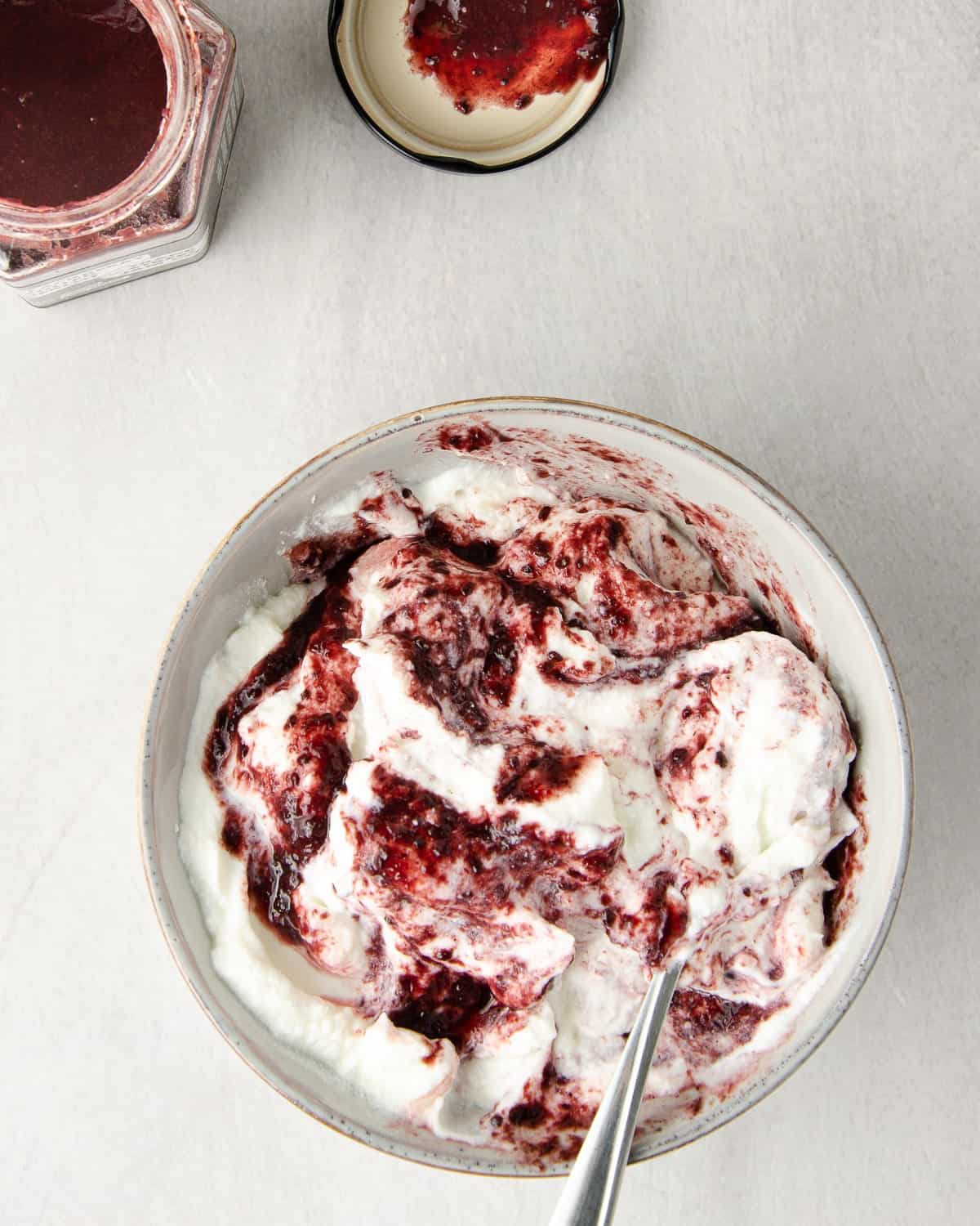 a center grey bowl of greek yogurt marbled with blackberry jam. the jam created swirls of a dark reddish color and is not fully mixed in. a jar of blackberry jam to the top left side