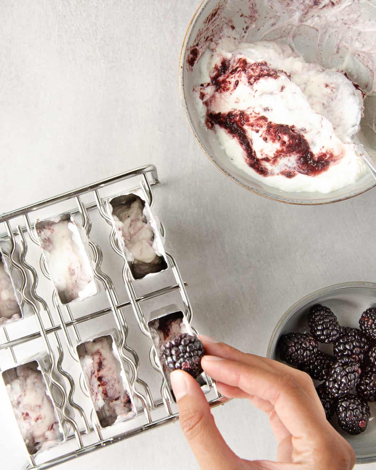placing a blackberry into half filled popsicle molds. a half-empty bowl of greek yogurt lightly marbled with blackberry jam