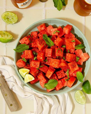 overhead view of a blue plate with watermelon tajin salad, some limes, a wooden knife and beers in the background