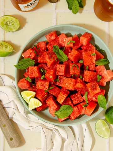 overhead view of a blue plate with watermelon tajin salad, some limes, a wooden knife and beers in the background