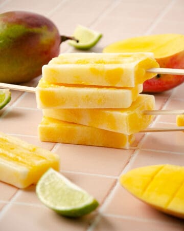 a stack of four mango popsicles with two mangos in the back, and sliced mango in the front, and lime wedges scattered around