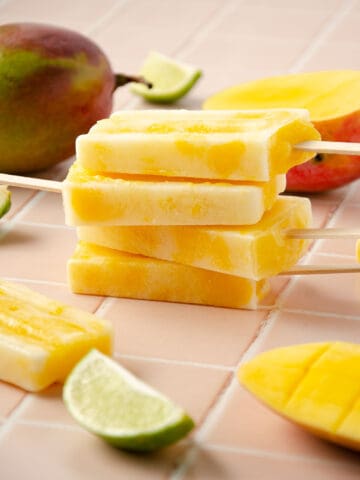 a stack of four mango popsicles with two mangos in the back, and sliced mango in the front, and lime wedges scattered around