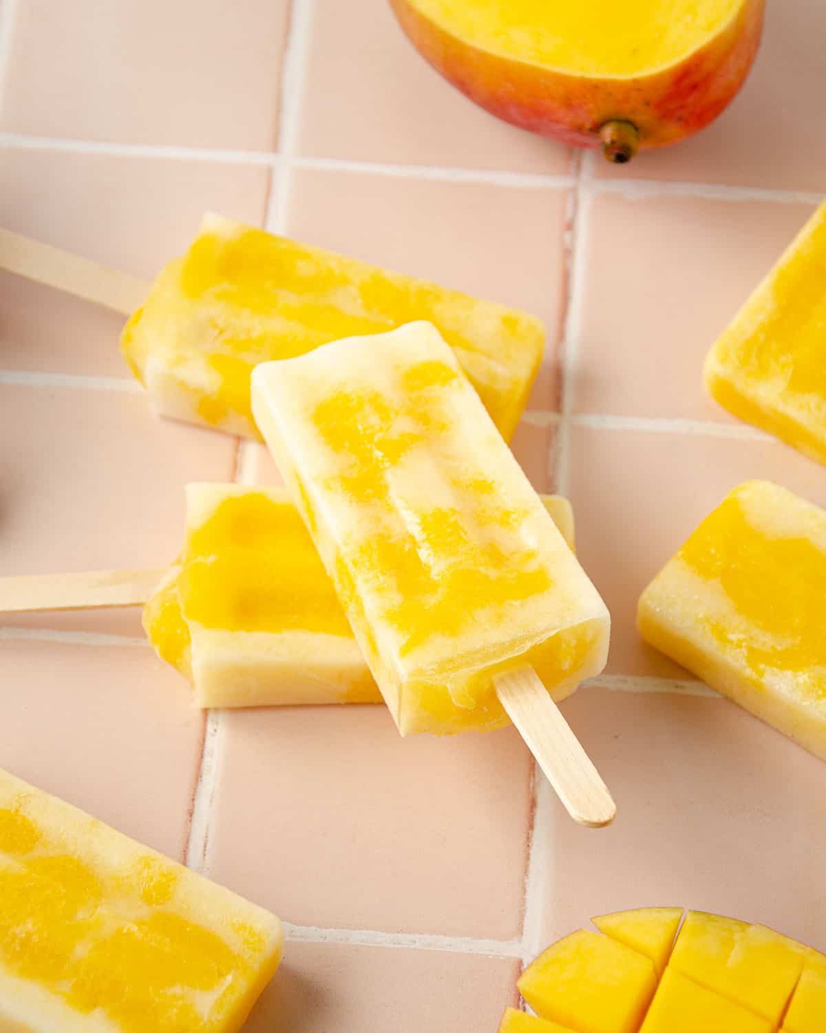 mango popsicles on top of one another on the pink countertop. two mango halves in the forefront of the photo