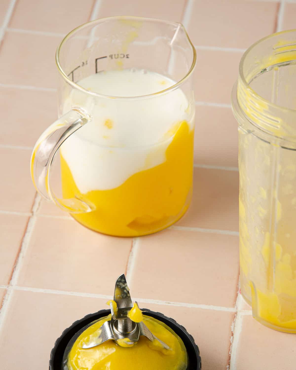 Mango Puree in a measuring cup with coconut milk on top of it. This is the step prior to mixing the milk and mango puree. A blender attachment with mango puree dripping off of it is in the front.