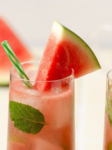 close up shot of a watermelon wedge sitting on a highball glass filled with a pink watermelon gin mojito with a light green striped straw and mint leaves