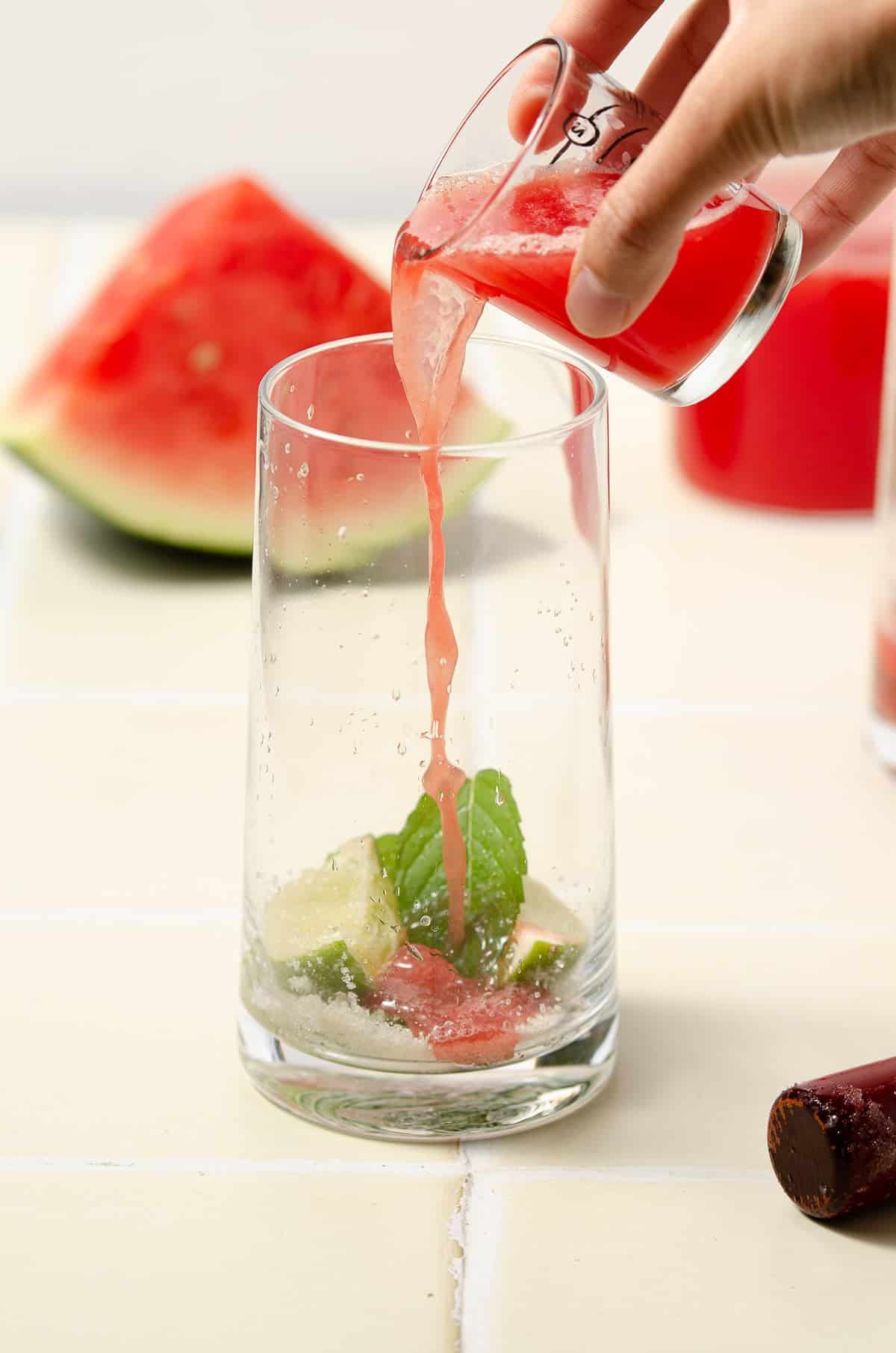 pouring watermelon juice on top of muddled lime wedges and mint leaves. a pitcher of watermelon juice and a watermelon wedge sit in the background