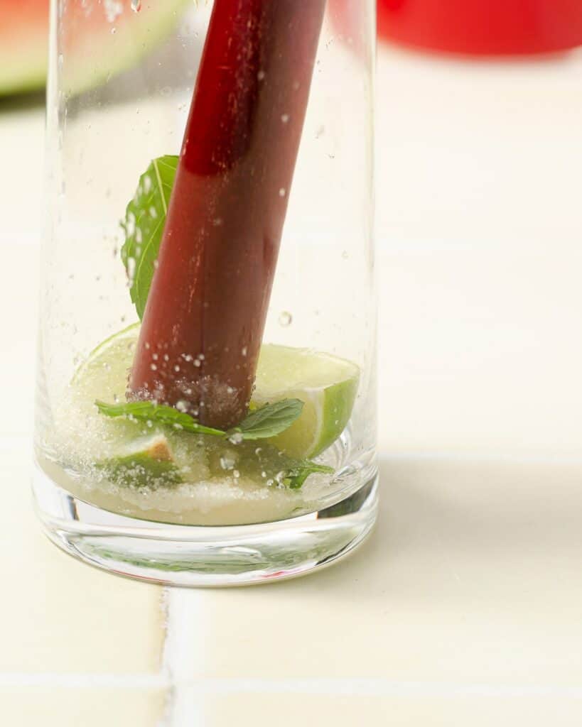 close up shot of a red wooden muddler muddling two lime wedges with mint leaves and granulated sugar
