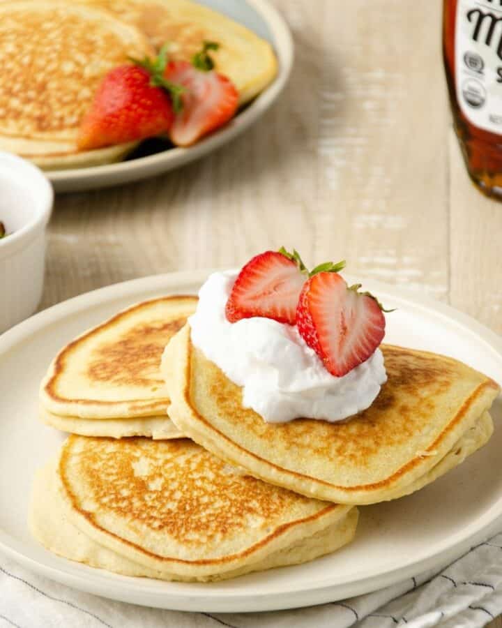 three oat milk pancakes layered on a plate topped with coconut whipped cream and halved strawberries. A small plate of pancakes sits to the back left with a small bottle of maple syrup sits behind.