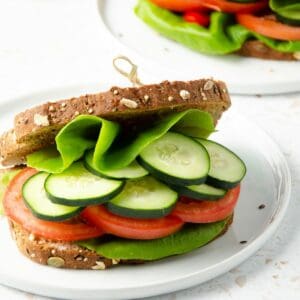 square feature photo of close up of a panera mediterranean veggie sandwich filled with butter lettuce, cucumber, tomato slices with a second sandwich and cutting board with fresh cucumber slices in the background.