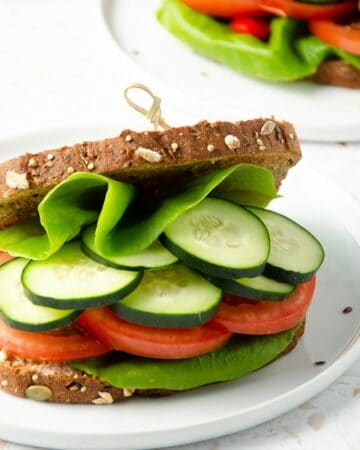 square feature photo of close up of a panera mediterranean veggie sandwich filled with butter lettuce, cucumber, tomato slices with a second sandwich and cutting board with fresh cucumber slices in the background.