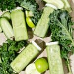 Featured Image of Four Glass bottles of green juice surrounded by chopped kale, green apples and cucumbers.