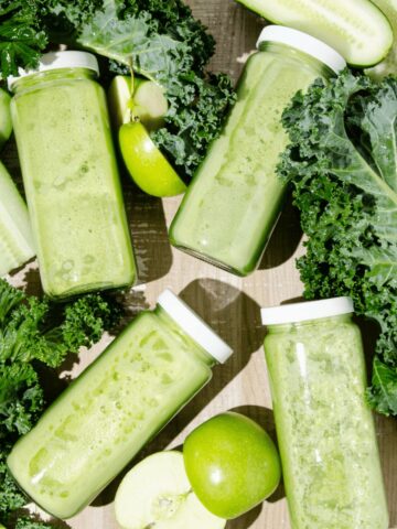 Featured Image of Four Glass bottles of green juice surrounded by chopped kale, green apples and cucumbers.