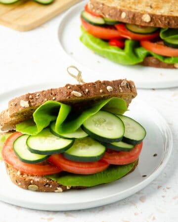 close up of a panera mediterranean veggie sandwich filled with butter lettuce, cucumber, tomato slices with a second sandwich and cutting board with fresh cucumber slices in the background.