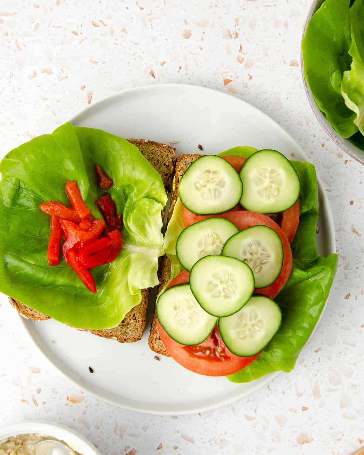 two slices of grain bread topped with butter lettuce and pimiento peppers on the left bread slice, and butter lettuce, tomato slices and cucumber slices on the right side. a bowl of butter lettuce and a bowl of mashed feta and hummus peeking on the sides.