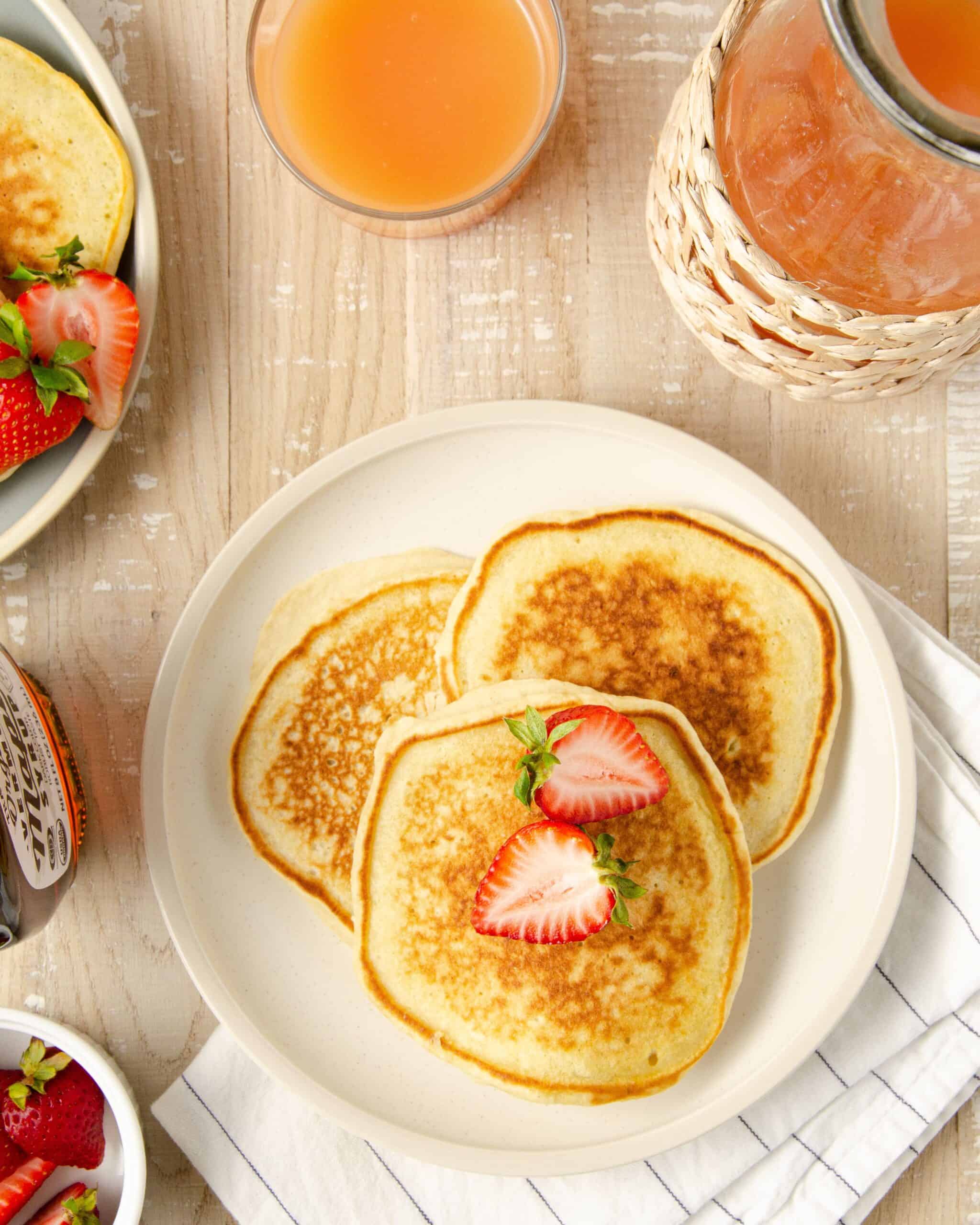 a beige plate of three pancakes topped with halved strawberry with a bowl of strawberries to the bottom, a second plate to the top left, and grapefruit juice