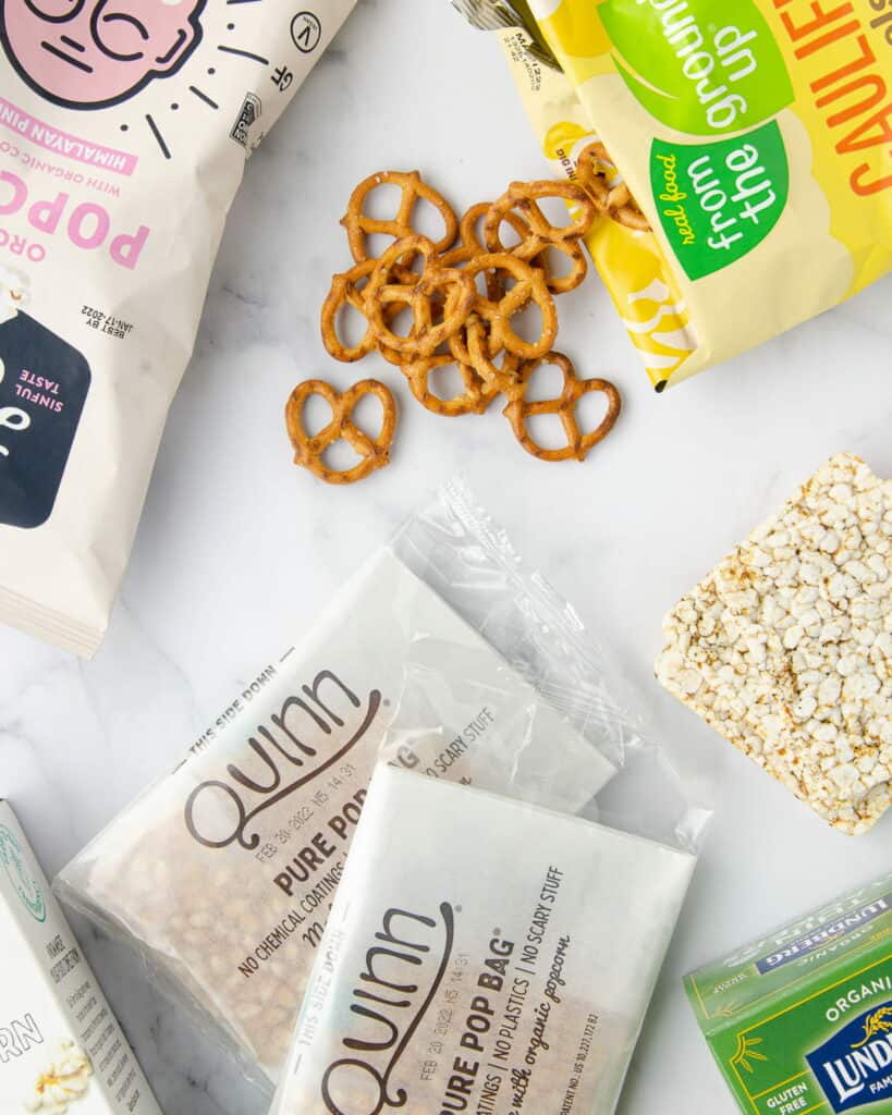 gluten free dairy free snacks on marble: featuring cauliflower pretzel twists, two bags of microwave popcorn, a bag of popped popcorn, and two rice cakes