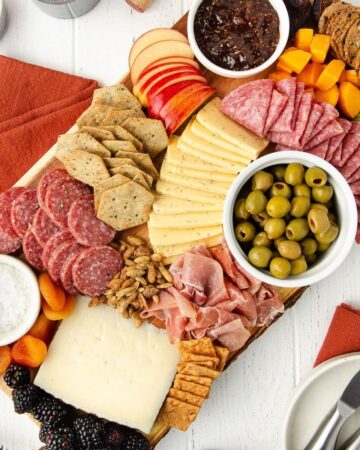 featured image of a gluten free charcuterie board sit-in on a white wooden table with orange linens and a plate with cheese knives sitting to the bottom