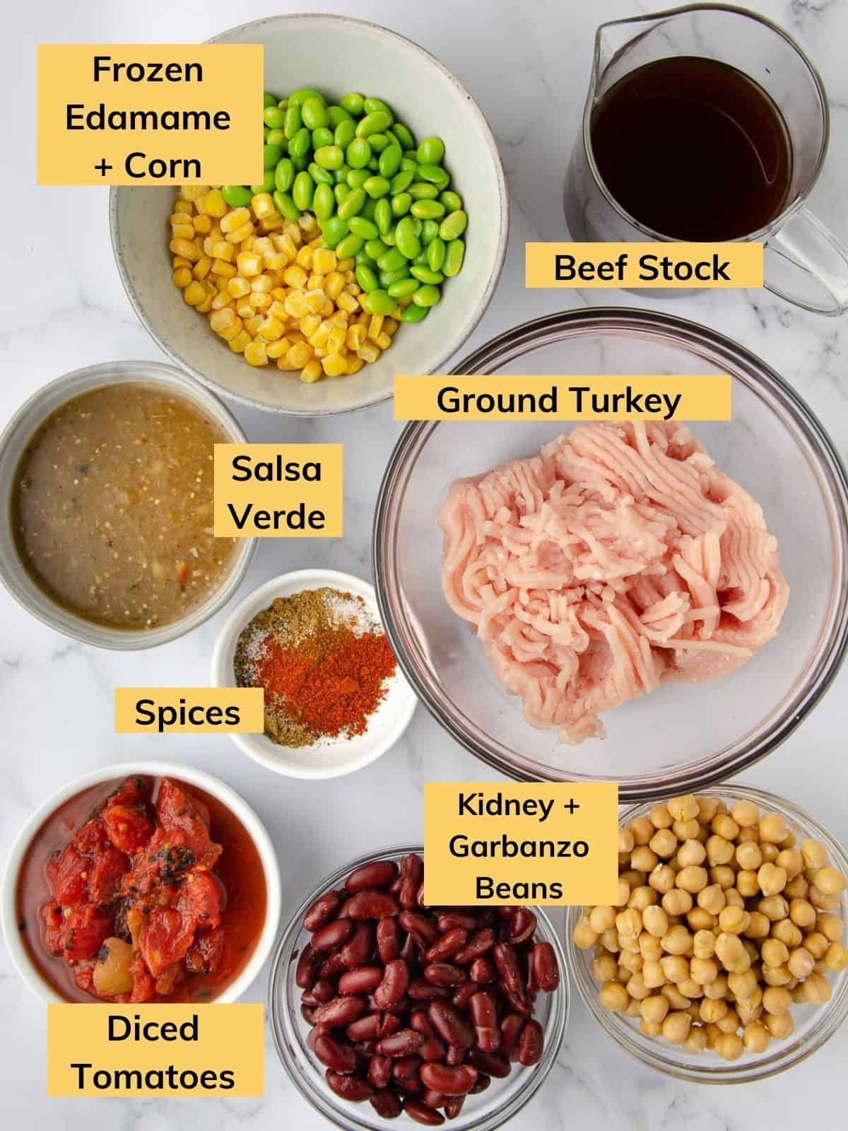 ingredients for panera turkey chili: beef stock, a bowl of ground turkey, a grey bowl of frozen edamame and corn, a bowl of salsa verde, a small bowl of spices, a bowl of diced tomatoes, and two bowls of drained beans.