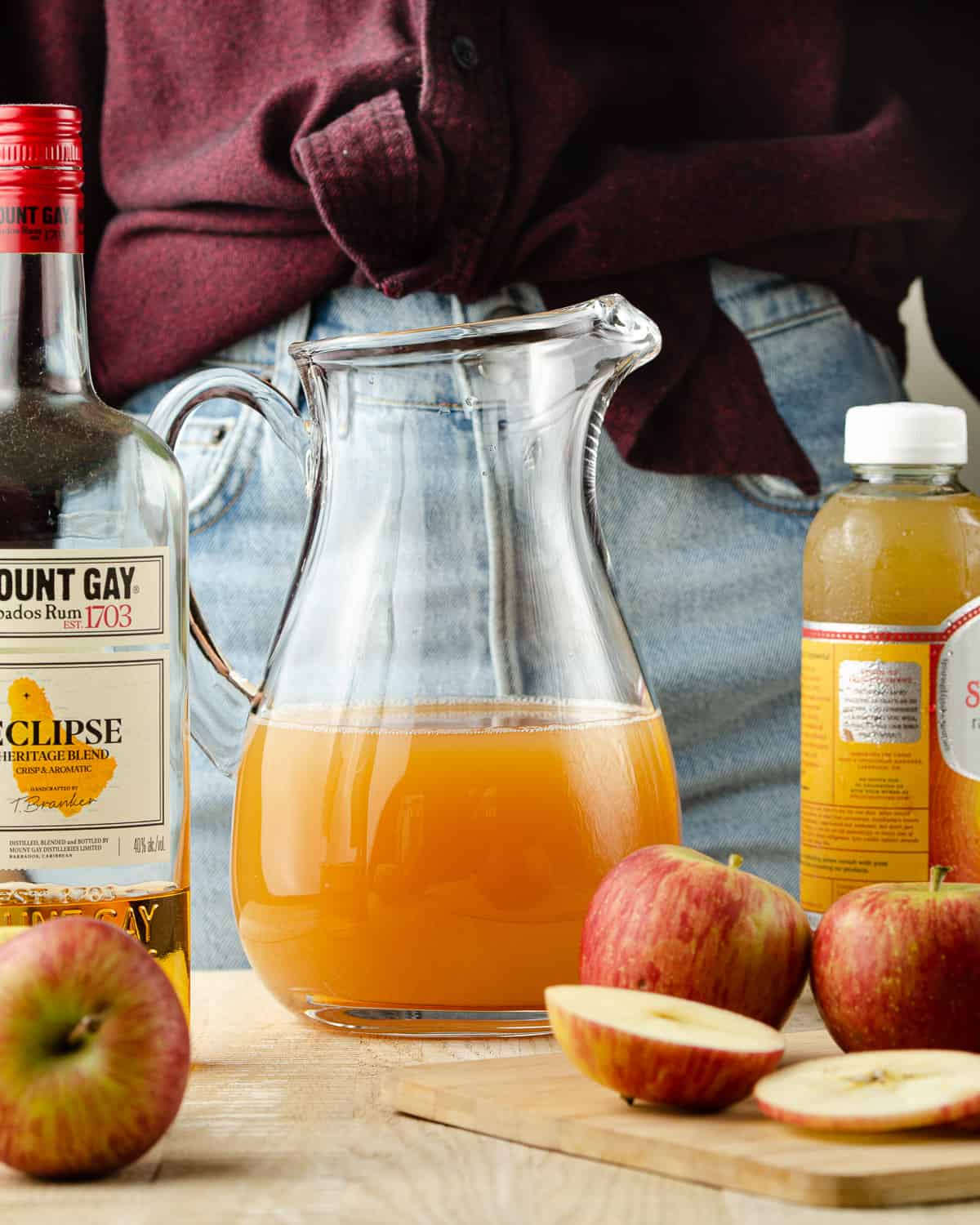 A pitcher of apple cider with a bottle of rum and a bottle of ginger kombucha to the side.